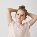 Close up of sweet little blonde girl with bun hairdo in pink t-shirt smiling, holding hands behind head with happy and satisfied expression.